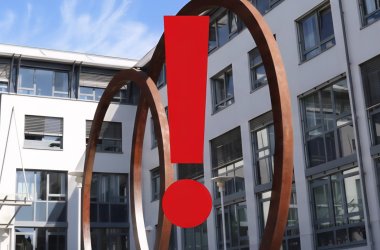 Ansicht des Jobcenters Pirmasens von außen, sichtbar ist die Frontansicht, davor eine große Skulptur aus zwei runden Kreisen. In der Mitte des Fotos befindet sich ein großes rotes Ausrufezeichen. 